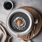 A bowl of creamy black sesame soup with a rich dark gray color and smooth, velvety texture, served in a white ceramic bowl, garnished with a few black sesame seeds on top and a drizzle of coconut milk creating an elegant swirl pattern, photographed from above on a clean wooden table with soft natural lighting, food photography style